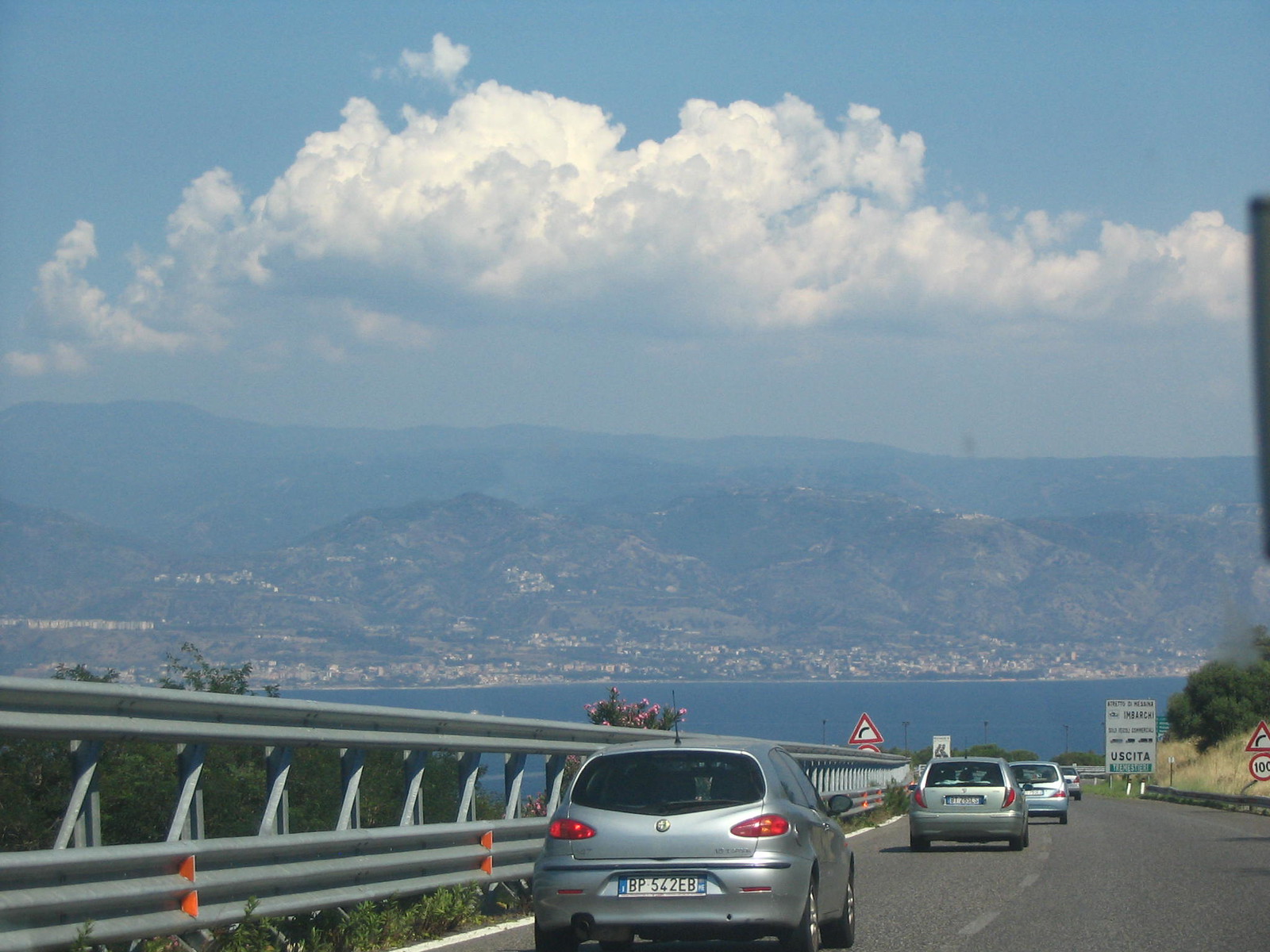 Auto in viaggio su una strada a scorrimento veloce vicino a Messina, con lo Stretto e la Calabria sullo sfondo
